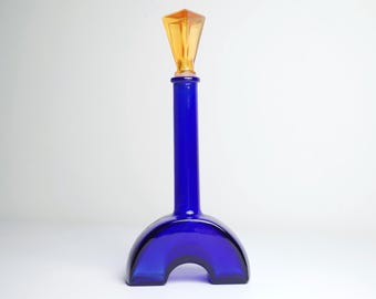 May include: A cobalt blue glass bottle with a unique arched base and a tall, slender neck. The bottle is topped with a faceted, amber-colored stopper. The design is modern and artistic.