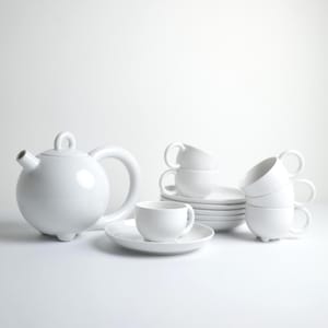 Może przedstawiać: Biały ceramiczny zestaw do herbaty, w tym okrągły czajniczek z zakrzywionym uchwytem, spodki i filiżanki. Filiżanki mają zaokrąglone uchwyty i stoją na pasujących spodkach. Zestaw jest prezentowany na białej powierzchni.