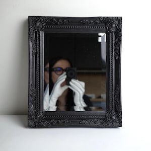 Peut inclure: Miroir rectangulaire noir avec un cadre orné et sculpté. Le cadre présente des motifs floraux et des volutes détaillés. Le miroir reflète une personne tenant un appareil photo. Un élément de décoration intérieure.
