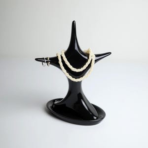 May include: A black jewelry display stand with a unique, star-like design. A cream-colored necklace and a pair of silver hoop earrings are displayed on the stand. The stand is made of a glossy material and has a small, oval base.