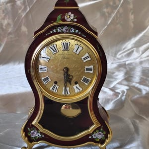Puede incluir: Un reloj de sobremesa color burdeos con esfera dorada, números romanos y el nombre Eluxa. La caja de madera presenta motivos florales pintados a mano, adornos dorados y un remate superior dorado. Un péndulo dorado es visible tras el panel de cristal.