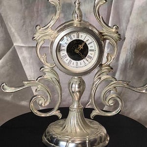 Può includere: Un orologio da tavolo in metallo color argento con una cornice ornata a volute e un quadrante circolare. Il quadrante ha uno sfondo bianco con numeri romani neri, un centro scuro e lancette sottili color oro su una base a piedistallo.