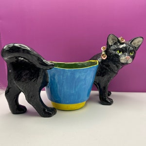 May include: Two black ceramic cat planter figurines. One cat is standing with a flower crown and the other is laying down with its tail curled around a blue and yellow planter.