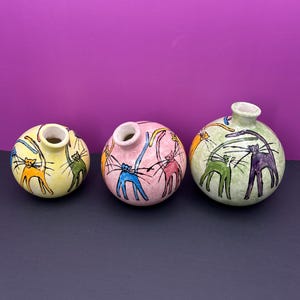 May include: Three ceramic vases with a round shape and a cat design. The vases are painted in different colors, including yellow, pink, and green. The cats are drawn in a whimsical style with colorful lines and details.