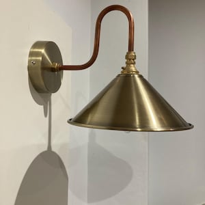 May include: A brass wall sconce with a copper pipe arm and a cone-shaped shade. The sconce has a simple design and a vintage aesthetic.