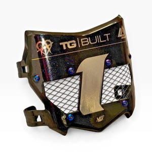 May include: A black motorcycle number plate with a glitter finish, featuring the text TG BUILT and a large number 1. The plate includes a silver metal mesh insert, blue hardware accents, and a small logo at the bottom right corner.