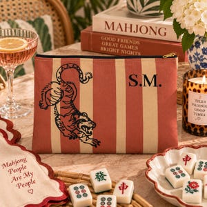 Può includere: Una pochette rettangolare con cerniera caratterizzata da un motivo a righe verticali color rosso tenue e crema. La parte anteriore mostra un'illustrazione a linee nere di una tigre e le lettere monogramma nere S.M. La pochette è in tessuto.