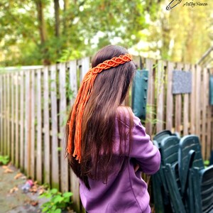 Puede incluir: Una mujer con el pelo largo y castaño lleva un jersey morado y pantalones negros. Lleva una diadema trenzada naranja con una larga borla.