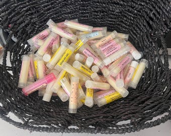 May include: A woven black basket overflowing with lip balm tubes. The tubes are clear, with pink, yellow, and white labels. Some labels have text. The lip balms are a light yellow color. The basket is made of thick, dark rope.