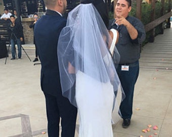 May include: A wedding ceremony taking place outdoors with the bride and groom standing under a white canopy. The bride is wearing a white wedding dress with a veil and the groom is wearing a dark suit. A person is standing behind the couple, possibly an officiant, and there are other guests in the background.