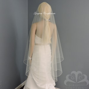 May include: A white single-tier wedding veil with a scalloped edge. The veil is made of soft tulle and is worn by a mannequin with blonde hair.