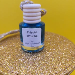 Könnte beinhalten: Kleine blaue Glasflasche mit Holzverschluss und weißem Etikett mit der Aufschrift "Frische Wäsche" und "8 ml". Die Flasche steht auf einer goldglitzernden Oberfläche. Der Hintergrund ist gelb.