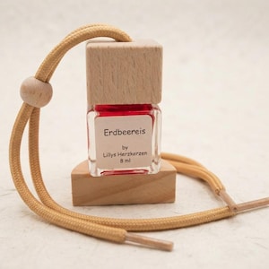 Könnte beinhalten: Ein kleiner quadratischer Glasflakon mit roter Flüssigkeit und einem hellen Holzdeckel. Das Etikett trägt die Aufschrift "Erdbeereis by Lillys Herzkerzen 8 ml". Eine beige Kordel mit Holzperle zum Aufhängen liegt auf einem Holzsockel.