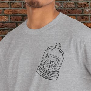 May include: A light grey t-shirt with a black and white graphic of a brain in a glass cloche. The text "DO NOT USE THIS BRAIN! - ABNORMAL" is printed below the cloche.