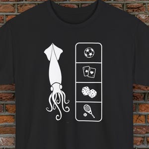 Può includere: T-shirt nera con grafica bianca. La grafica presenta un calamaro a sinistra e un rettangolo verticale a destra con un pallone da calcio, carte da gioco, dadi e una racchetta da tennis.