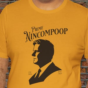 Może przedstawiać: Musztardowa koszulka z czarną grafiką przedstawiającą mężczyznę w garniturze i napisem "Prime Nincompoop" nad obrazem. Koszulka zawiera również tekst "Est. 2024" i "Diss. 2029".