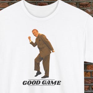 May include: White t-shirt with a printed image of a man in a brown suit, celebrating. Below the image, the text reads "GOOD GAME" in black. A humorous, novelty t-shirt.