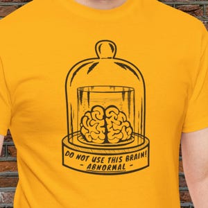 May include: Mustard yellow t-shirt with a black and white graphic of a brain in a glass case. The text reads "DO NOT USE THIS BRAIN! - ABNORMAL - ".