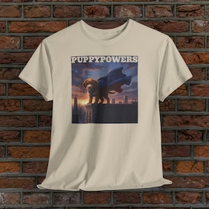 May include: Beige t-shirt featuring a graphic of a puppy wearing a cape, standing on a rooftop with a cityscape backdrop. The text "PUPPYPOWERS" is above the image. The puppy is brown with a blue cape.