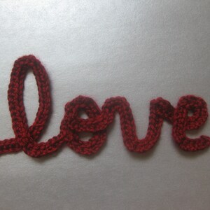 Peut inclure: Mot crocheté rouge formant le mot "love" sur fond gris. Le fil est d'un rouge profond, avec des points visibles créant un aspect texturé. Le mot est écrit dans un style cursif.