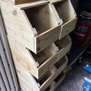 Puede incluir: Unidad de almacenamiento de madera con cuatro compartimentos inclinados, construida con madera sin tratar. Cada compartimento es accesible desde el frente. Tablas de madera adicionales apiladas en la parte superior.