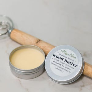 May include: A small, open metal tin of wood butter with a light yellow color, next to its lid. The lid has text that reads "May Farm wood butter" and instructions. A wooden whisk is in the background.