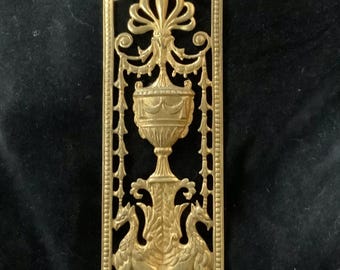 May include: A gold metal decorative plate with an ornate design featuring a vase with a floral motif and two stylized animals. The plate has a rectangular shape and a detailed border.