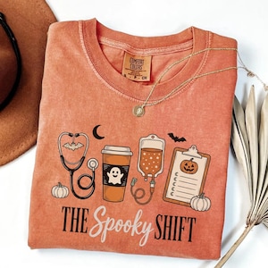 May include: A burnt orange short-sleeve t-shirt featuring a graphic print of a stethoscope, a coffee cup with a ghost, an IV bag with stars, and a clipboard with a pumpkin. The text reads THE SPOOKY SHIFT. The shirt has a Comfort Colors brand label.