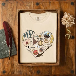 May include: A cream-colored t-shirt featuring a heart-shaped graphic composed of various illustrations including a castle, a sorting hat, a wand, a golden snitch, glasses, a broomstick, a letter, and text reading HP, Muggles, 9 3/4, Always, and it's leviOsa, not leviosA.
