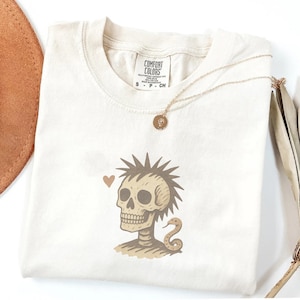May include: An off-white crew neck t-shirt with a Comfort Colors label. The graphic features a tan skull with a spiky mohawk hairstyle, a small brown heart, and a coiled snake. A thin gold-toned necklace with a small circular pendant rests on the shirt.