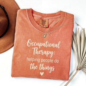 May include: A terracotta-colored short-sleeve t-shirt with a crew neckline. The shirt features the white printed text Occupational Therapy: helping people do the things and a small heart icon. A gold-toned necklace rests on the fabric.