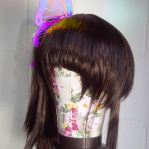 May include: A brown wig with a iridescent butterfly hair clip. The butterfly is purple, pink, and blue.