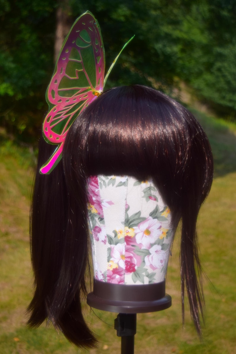May include: A dark brown wig with long straight hair and bangs. The wig is on a floral-patterned mannequin head and has a pink and green butterfly hair clip.