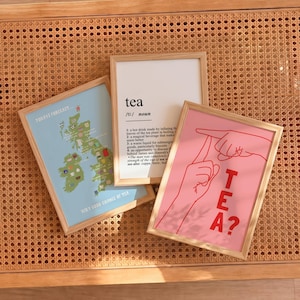 May include: Three framed wall art prints. One print features a light blue map of the British Isles with tea-themed icons. Another is a white dictionary-style definition of tea. The third is a pink print with red hands and the text TEA?