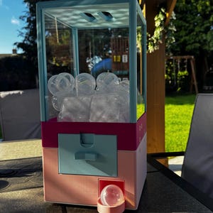 May include: A vertical claw machine toy with a light blue frame, a magenta middle section, and a light pink base. The top section is clear plastic filled with empty transparent capsules, and a single capsule sits in the light pink dispensing tray.