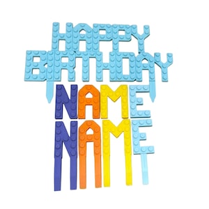May include: A blue and light blue cake topper with the words "HAPPY BIRTHDAY" and "NAME" in a block-style design. The letters are in blue, orange, and yellow. The topper is designed to be inserted into a cake.