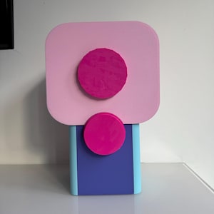 Op de afbeelding: Een moderne, abstracte sculptuur in roze, blauw en paars. De sculptuur heeft een rechthoekige basis in blauw en lichtblauw, bekroond met een roze vierkant en twee magenta cirkels. Het ontwerp is geometrisch en minimalistisch.