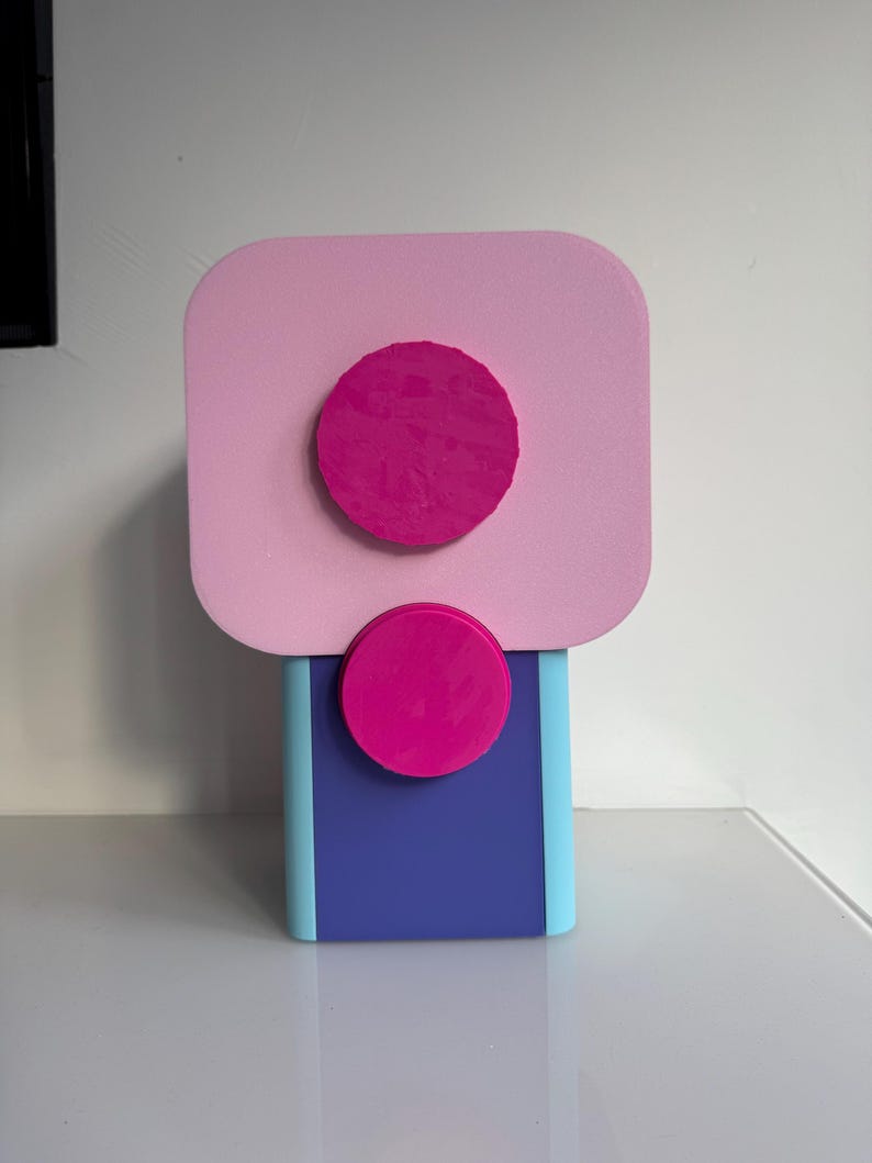 Op de afbeelding: Een moderne, abstracte sculptuur in roze, blauw en paars. De sculptuur heeft een rechthoekige basis in blauw en lichtblauw, bekroond met een roze vierkant en twee magenta cirkels. Het ontwerp is geometrisch en minimalistisch.