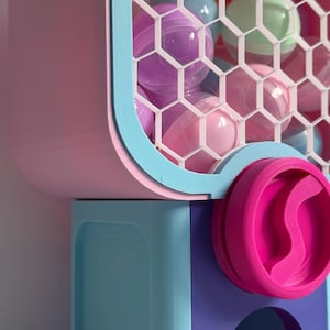 Op de afbeelding: Een kleurrijke kauwgomballenautomaat in roze, blauw en paars. De machine heeft een raam met honingraatpatroon gevuld met pastelkleurige kauwgomballen. Een roze doseerwiel en een bakje maken het ontwerp compleet.