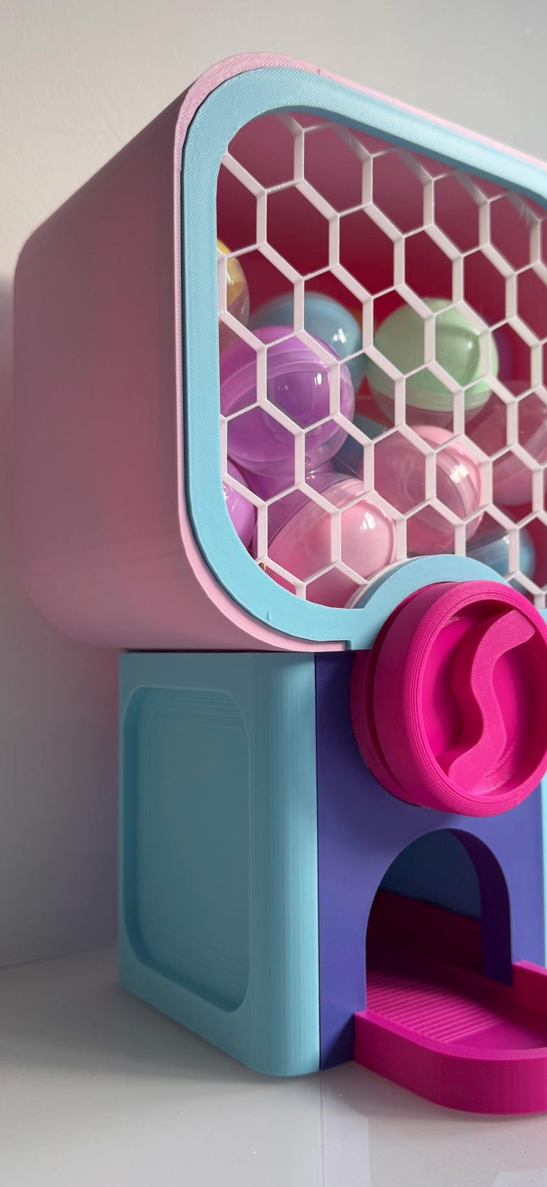Op de afbeelding: Een kleurrijke kauwgomballenautomaat in roze, blauw en paars. De machine heeft een raam met honingraatpatroon gevuld met pastelkleurige kauwgomballen. Een roze doseerwiel en een bakje maken het ontwerp compleet.