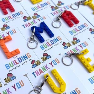 May include: Keychains shaped like the letters A, E, L, M, and U, made from colorful plastic blocks. Each keychain has a silver keyring attached. The keychains are displayed on cards that say "Thank You" and "Building memories with me."