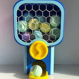 Op de afbeelding: Een kleurrijke kauwgomballenautomaat in blauw en geel, met een honingraatpatroon venster gevuld met verschillende gekleurde ballen. Een enkele bal rust in de uitgiftebak. De machine heeft een muntsleuf met een gele 'S'.