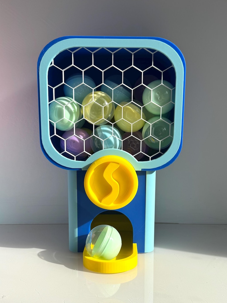 Op de afbeelding: Een kleurrijke kauwgomballenautomaat in blauw en geel, met een honingraatpatroon venster gevuld met verschillende gekleurde ballen. Een enkele bal rust in de uitgiftebak. De machine heeft een muntsleuf met een gele 'S'.