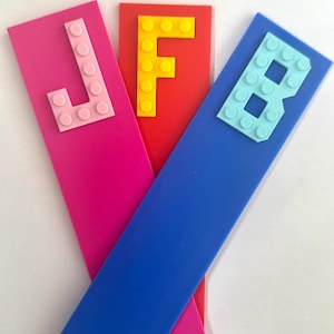 Può includere: Tre segnalibri colorati con lettere. Un segnalibro rosa ha la lettera "J" rosa chiaro fatta di mattoncini giocattolo. Un segnalibro rosso ha la lettera "F" gialla. Un segnalibro blu ha la lettera "B" azzurra.