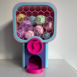 Op de afbeelding: Een kleurrijke kauwgomballenautomaat met een roze, blauw en paars ontwerp. Het bovenste gedeelte bevat meerdere doorzichtige plastic ballen in verschillende pastelkleuren. De machine heeft een uitgiftesleuf en een ronde roze draaihendel.