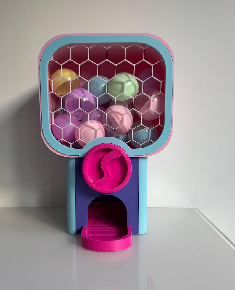 Op de afbeelding: Een kleurrijke kauwgomballenautomaat met een roze, blauw en paars ontwerp. Het bovenste gedeelte bevat meerdere doorzichtige plastic ballen in verschillende pastelkleuren. De machine heeft een uitgiftesleuf en een ronde roze draaihendel.