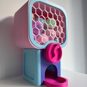 Op de afbeelding: Een kleurrijke kauwgomballenautomaat in roze, blauw en paars. De machine heeft een doorzichtig raam met honingraatpatroon, gevuld met verschillende gekleurde kauwgomballen. Een roze hendel en bakje zijn zichtbaar.