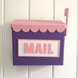 以下が含まれることがあります： ピンクのスカラップ屋根とピンクの旗が付いた紫色の郵便受け。「MAIL」の文字がピンク色の文字で白い長方形の背景に表示されています。郵便受けは、白い垂直パネルの表面に取り付けられています。