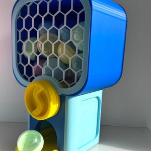 Op de afbeelding: Een blauw en gele kauwgomballenautomaat met een honingraatfront. De machine geeft transparante, kleurrijke kauwgomballen af. Het ontwerp heeft een felblauwe bovenkant, een lichtblauwe basis en gele accenten.