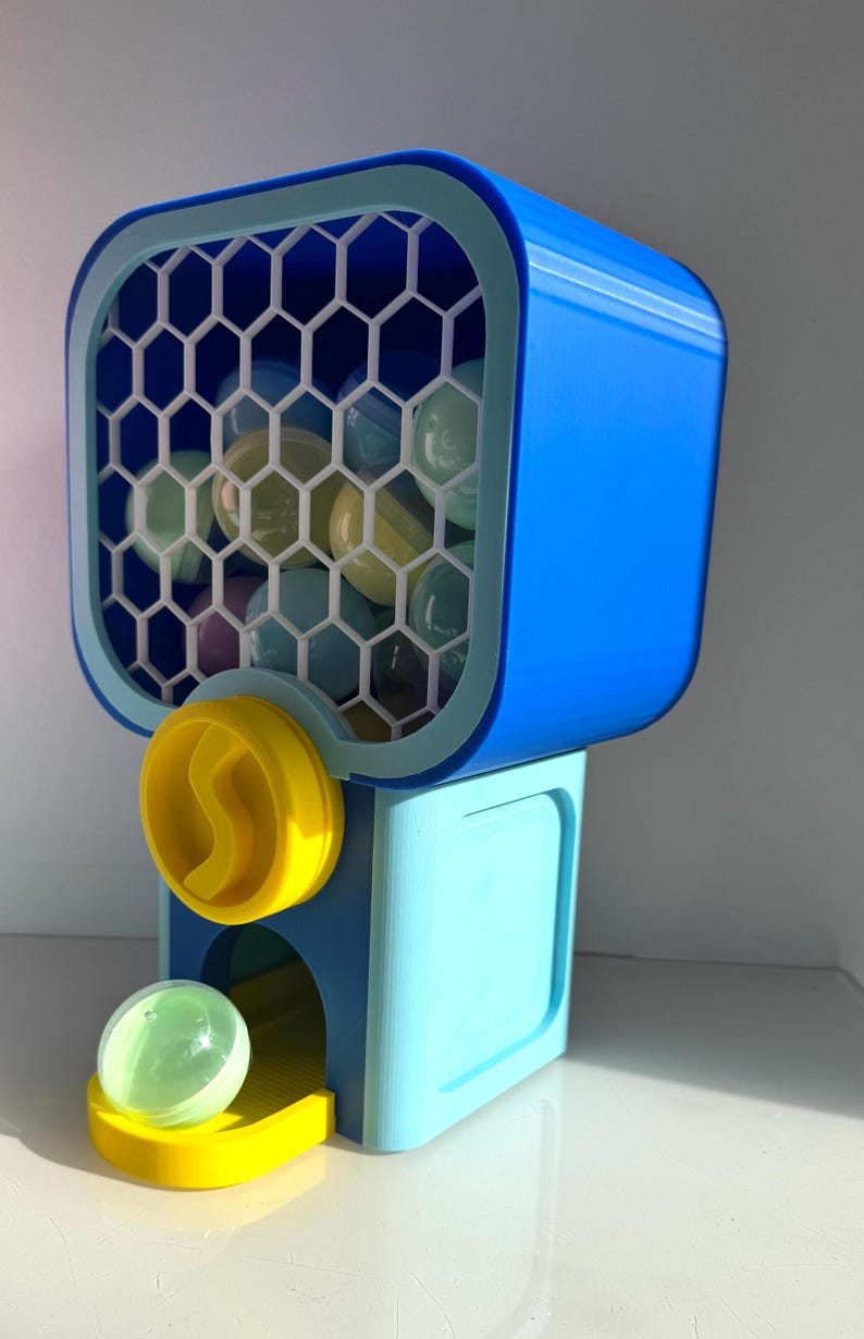 Op de afbeelding: Een blauw en gele kauwgomballenautomaat met een honingraatfront. De machine geeft transparante, kleurrijke kauwgomballen af. Het ontwerp heeft een felblauwe bovenkant, een lichtblauwe basis en gele accenten.